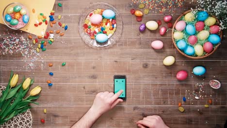Man-using-smartphone-with-green-screen-on-table-decorated-with-easter-eggs.-Top-view