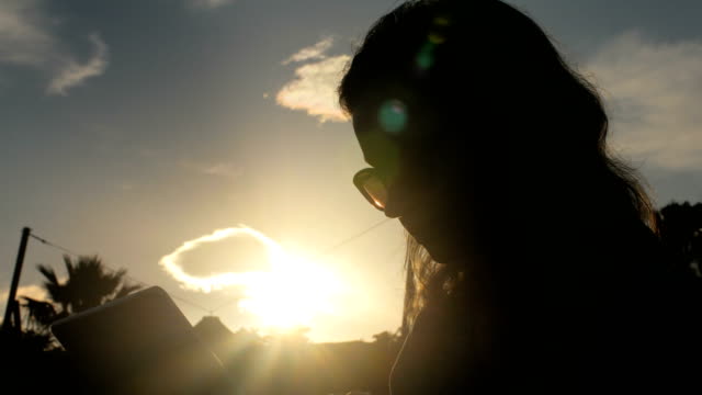 young-woman-with-sunglasses-using-tablet,-against-sun-shot,-clouds-and-silhouette-of-palms