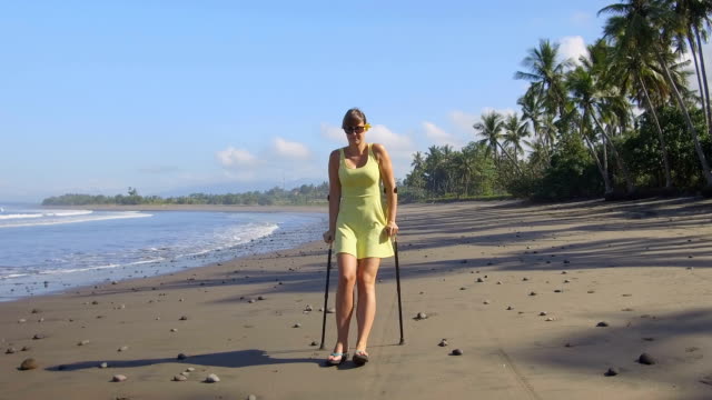 CLOSE-UP-Woman-with-crutches-walking-along-the-coastline-on-tropical-island-Bali