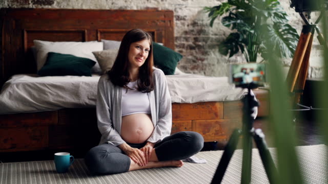 Schöne-junge-Frau-Blogger-ist-Video-über-Schwangerschaft-für-ihr-Vlog-zu-Hause-auf-dem-Boden-sitzen,-reden-und-Blick-in-die-Smartphone-Kamera-auf-Stativ-aufgenommen.