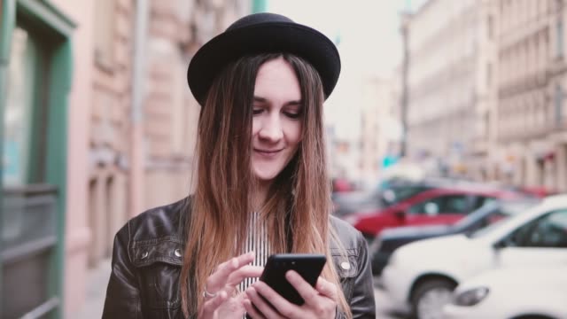 Lächelnde-junge-Europäerin-in-stilvollen-Hut-entlang-Stadtstraße-mit-einem-Smartphone-Zeitlupe-vorwärts-schauen
