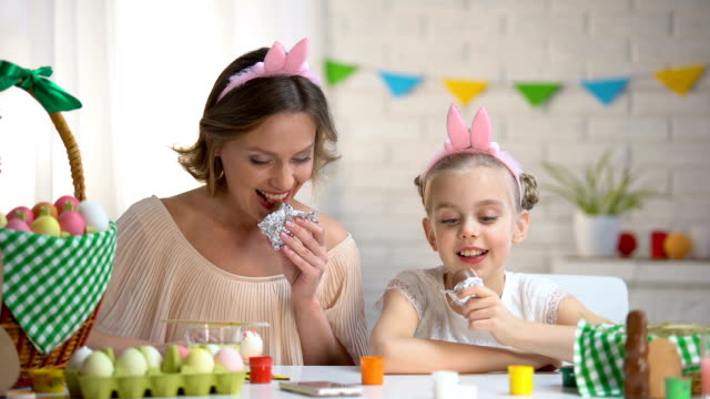 Schöne-Mutter-und-Tochter-in-lustige-Stirnbänder-essen-Schokolade-Ostereier
