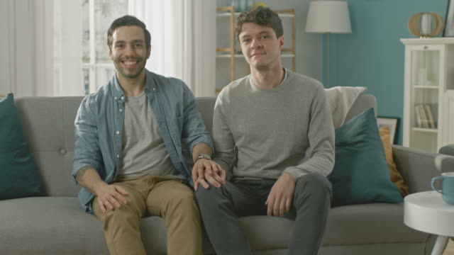 Cute-Attractive-Male-Gay-Couple-Sit-Together-on-a-Sofa-at-Home.-Boyfriend-Puts-His-Hand-on-Fiance's.-They-are-Happy-and-Smiling.-They-are-Casually-Dressed-and-Their-Room-Has-Modern-Interior.
