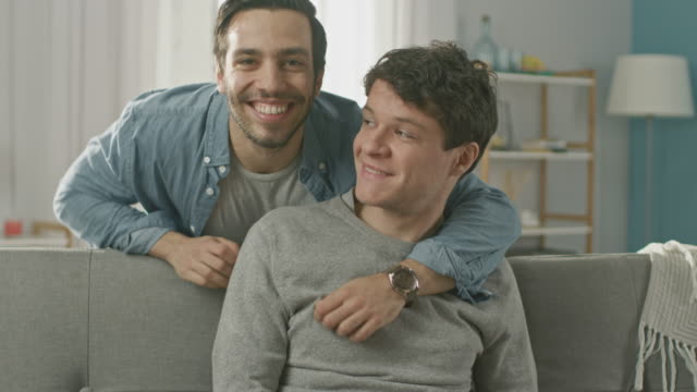 Portrait-of-a-Cute-Male-Queer-Couple-at-Home.-Young-Man-Sits-on-a-Couch,-His-Partner-Comes-From-Behind-and-Gently-Embraces-Him.-They-are-Happy-and-Smiling.-Room-Has-Modern-Interior.