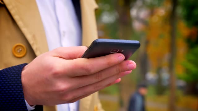Closeup-of-male-hand-holding-modern-smartphone-and-managing-it-on-the-background-of-nature-outdoors