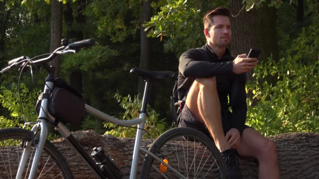 Ein-junger-hübscher-Radfahrer-sitzt-auf-einem-Balken-neben-seinem-Fahrrad-in-einem-Wald-und-arbeitet-auf-einem-Smartphone