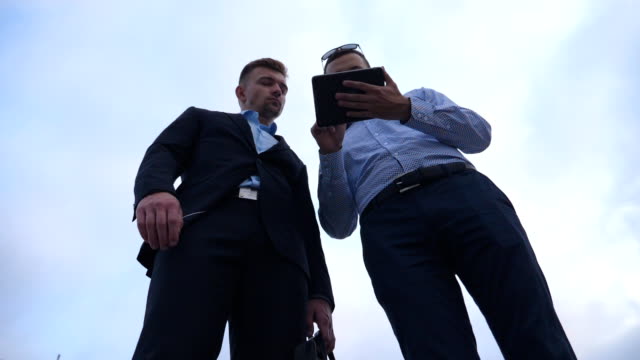 Low-angle-view-of-young-businessman-showing-a-presentation-on-screen-of-tablet-pc-to-his-colleague-in-city.-Successful-entrepreneurs-working-on-digital-tablet.-Blue-sky-at-background.-Slow-motion