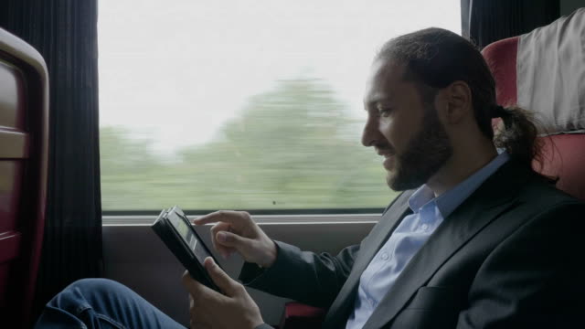 Feliz-joven-empresario-hombre-conmutando-en-tren-relajante-su-viaje-de-negocios-viendo-a-través-de-la-ventana-y-navegar-por-Internet-en-Tablet-PC