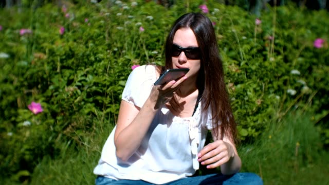 Beautiful-brunette-woman-records-a-voice-message-on-her-mobile-phone-while-sitting-in-the-park-on-a-Sunny-day.
