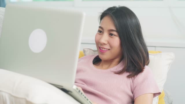 Young-business-freelance-Asian-woman-working-on-laptop-checking-social-media-while-lying-on-the-sofa-when-relax-in-living-room-at-home.-Lifestyle-latin-and-hispanic-ethnicity-women-at-house-concept.