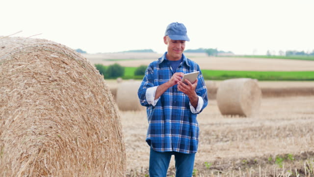 Farmer-using-digital-tablet--computer-while-standing-by-hay-bale.-Modertn-Agriculture