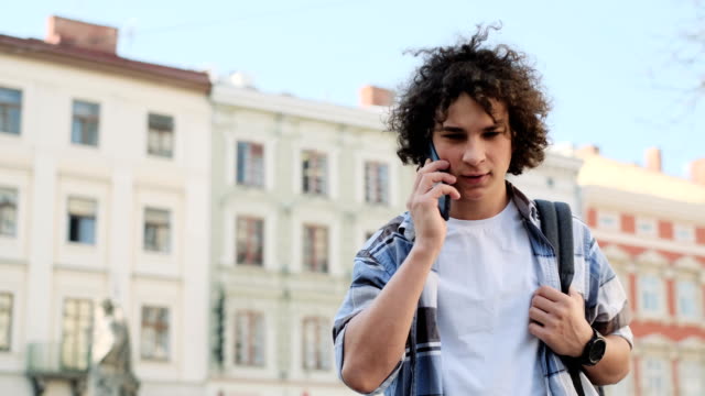 A-man-is-talking-on-the-phone-and-smiling.-Portrait-of-handsome-blogger,-student-walking-outdoors-and-talking-on-mobile-smartphone.-Education,-travel,-tourism-concept