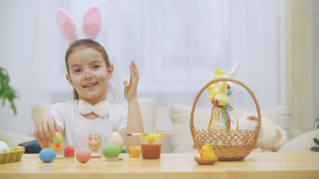 Little-cute-and-adorable-girl-is-smiling-sincerely.-She-takes-an-Easter-egg-and-shows-the-result-of-her-work,-using-her-dirty-of-paints-hands.-Girl-is-waving.