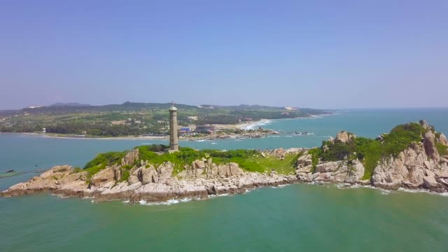 Sea-light-house-on-rocky-island-in-blue-sea-and-modern-city-on-skyline,-aerial-landscape-from-drone.-Scenic-view-lighthouse-tower-on-green-island-in-ocean