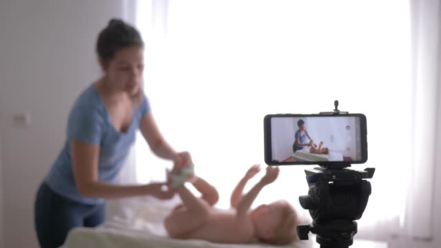 educational-video-blog-for-young-moms,-young-mom-vlogger-changes-diaper-to-small-child-boy-while-recording-education-video-for-followers-live-on-smart-phone-indoors