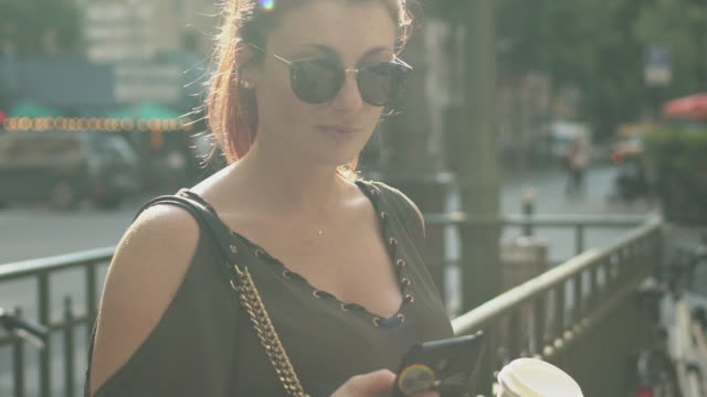 Young-attractive-woman-writing-a-text-message-on-her-smartphone-at-the-subway-exit-in-street,-holding-her-coffee-in-other-hand,-during-sunny-summer-afternoon-in-Paris.-Trendy-and-cool,-slow-motion.