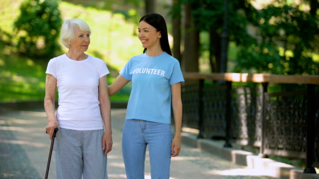 Female-volunteer-and-senior-patient-with-stick-walking-hospital-garden,-care