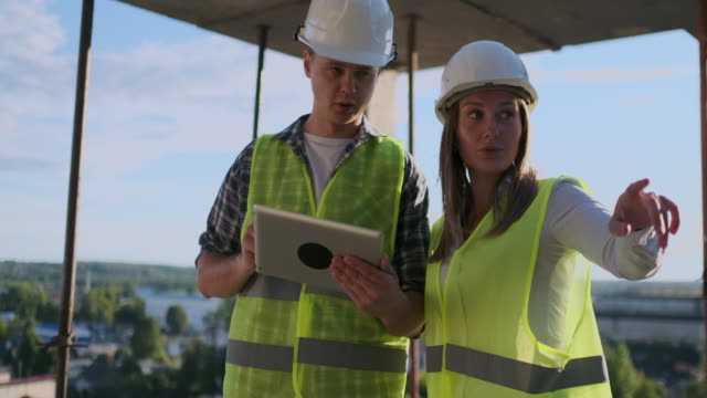 Construction-worker-man-and-architect-woman-in-a-helmet,-discuss-the-plan-of-construction-of-house,-tell-each-other-about-the-design,-holding-a-tablet,-look-at-the-drawings,-background-of-sun-rays
