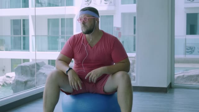 Funny-fat-male-in-pink-glasses-and-in-a-pink-t-shirt-is-engaged-on-a-fit-ball-in-the-gym-depicting-a-girl.-A-wonderful-man-listens-to-music-and-dances-on-the-ball-in-the-gym