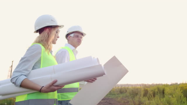 Ein-Mann-und-eine-Baufrau-mit-Zeichnungen-in-weißen-Helmen-gehen-und-reden-auf-der-Baustelle