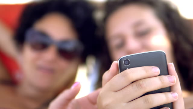 Teens-Lächeln-Vorschau-Social-Media-Posts-auf-Smartphone-liegend-auf-Strandbett,-selektiver-Fokus