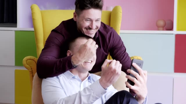 Gay-couple-using-phone-on-arm-chair-together.-Showing-ring-via-video-call.
