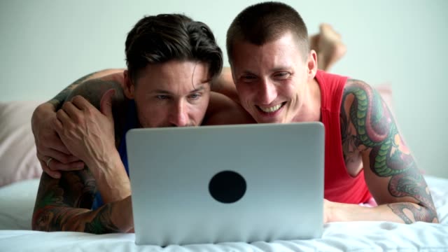 Pareja-gay-en-la-cama-usando-computadora-portátil.-Abrazando-y-sonriendo.