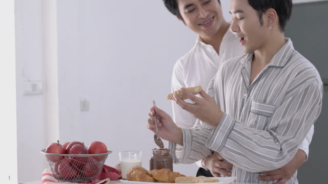 Asian-couple-gay-applying-chocolate-on-bread-in-morning-at-home.-Gay-boy-happy-emotion-and-hug-love-anniversary-together.-Concept-of-lifestyle,-family,-gay-and-bisexual.