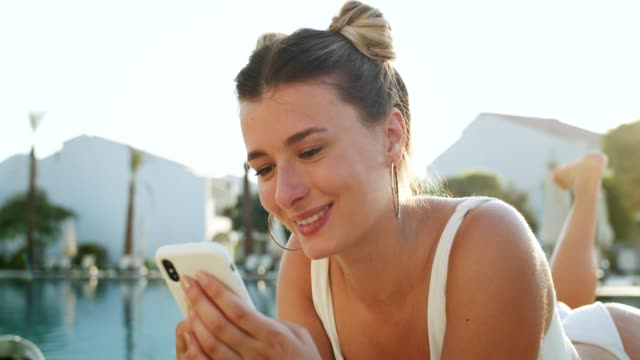 Close-up-of-the-pretty-Caucasian-young-girl-sunbathing-at-the-swimming-pool-and-using-her-smartphone-for-playing-or-social-media,-then-smiling-happily-to-the-camera.-Portrait.
