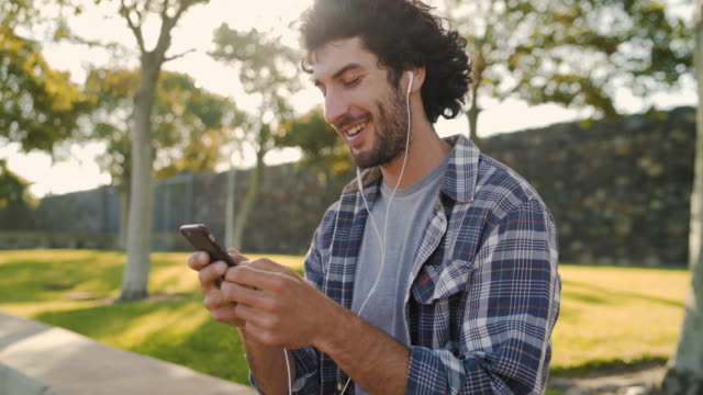 Primer-plano-de-un-joven-feliz-sonriente-con-auriculares-blancos-en-los-oídos-usando-el-móvil-para-mensajes-de-texto-en-el-parque