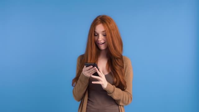Redheaded-girl-in-brown-t-shirt-and-cardigan.-She-is-using-smartphone,-smiling-and-looking-surprised-while-posing-against-blue-background.-Close-up