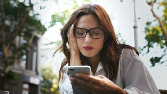 Business-woman-in-glasses,-white-shirt.-She-feeling-disappointed-while-reading-news-using-smartphone,-sitting-in-outdoor-cafe.-Close-up,-slow-motion