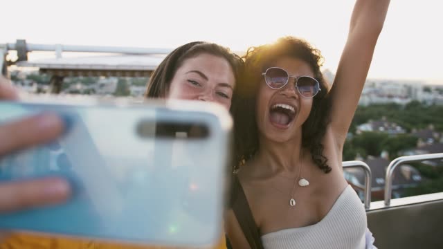 Frauen-zeigen-Zungen,-lächeln-und-machen-Selfie-auf-dem-Smartphone,-während-sie-in-einer-Riesenradkabine-sitzen.-Sonniger-Tag-im-Park.-Nahaufnahme,-Zeitlupe