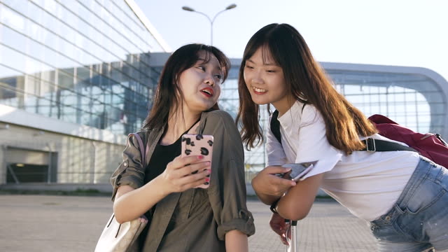 Charming-cheerful-25-years-old-asian-female-travelers-standing-near-modern-airport-and-using-their-smartphones