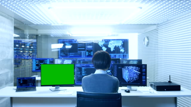 Back-View-of-the-IT-Technician-Working-on-with-Neural-Network-on-His-Personal-Computer-with-Green-Screen-Mock-up.-He-Works-in-a-Big-System-Control-Data-Center-with-Multiple-Monitors-Showing-Graphics.