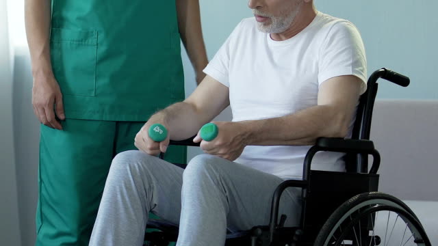 Aged-man-sitting-in-wheelchair,-holding-dumbbells,-talking-to-nurse,-recovery