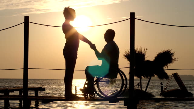 silhouette-of-female-and-male-handicapped-on-wheel-chair-holding-arms-and-look-at-horizon-in-sea