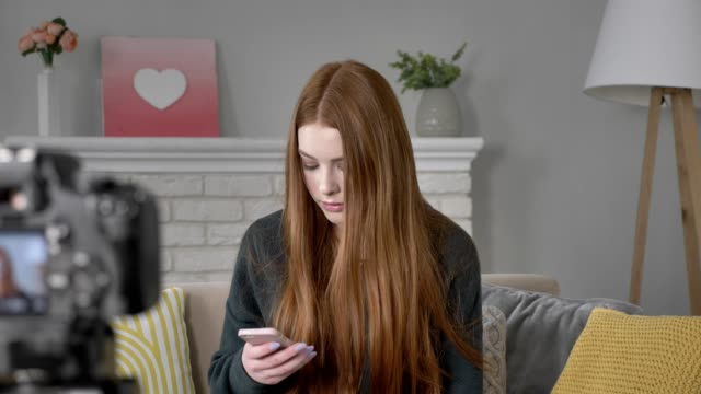 Young-red-haired-girl-blogger-at-the-camera,-using-a-smartphone,-texting,-typing,-home-cosiness-in-the-background.-60-fps