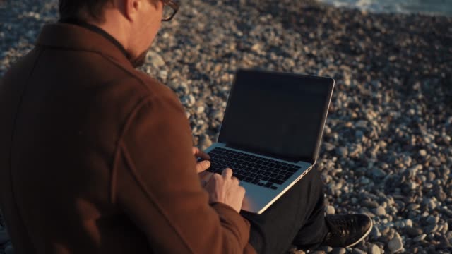 Mann-arbeitet-mit-Laptop-sitzen-Kieselstrand-von-Meer-abends-an