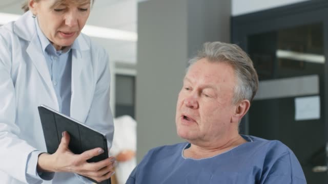 In-the-Hospital-Female-Doctor-Shows-Tablet-Computer-to-Elderly-Patient,-Explaining-his-Condition.-Modern-Hospital-with-Best-Possible-Care.