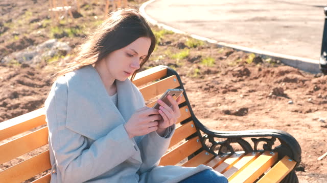 Portrait-of-beautiful-brunette-reading-and-typing-a-message-in-her-phone-sitting-on-the-bench-in-park.