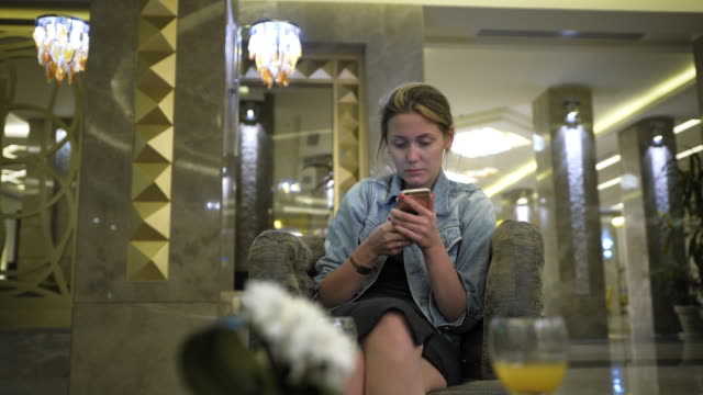 young-Caucasian-woman-in-a-jeans-jacket-uses-a-smartphone,-writes-messages-on-the-social-network.-Sits-at-the-table-in-the-hotel-restaurant.-bottom-view