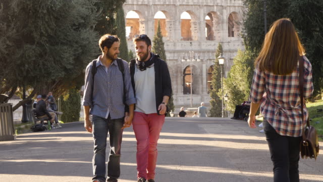Young-happy-gay-couple-tourists-walk-in-park-road-with-trees-colosseum-in-background-in-rome-at-sunset-holding-hands-gelousy-when-beautiful-attractive-girl-walks-by-glancing-lovely-slow-motion-colle-oppio