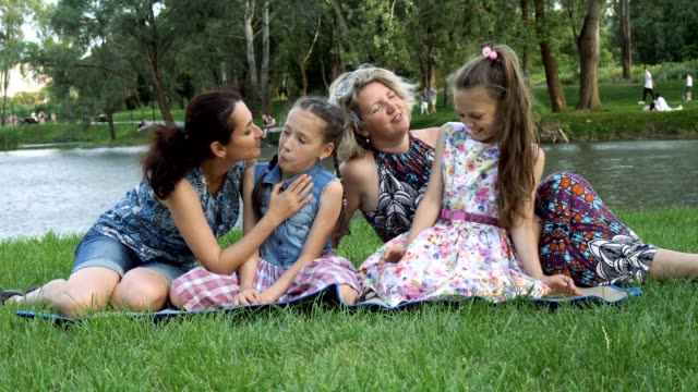 Glückliche-Homo-Familie:-zwei-Mütter-und-zwei-Töchter,-sitzen,-spielen-und-Lachen-auf-dem-Rasen-im-Park-am-Fluss-bei-Sonnenuntergang.
