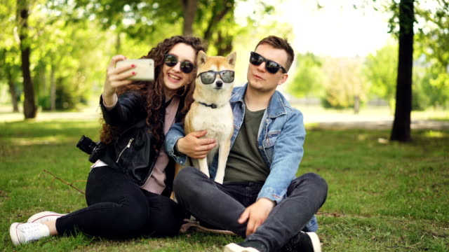 Pretty-woman-is-taking-selfie-with-her-boyfriend-and-adorable-dog-using-smartphone-while-resting-in-the-park-on-the-grass.-Humans-and-animals-are-wearing-sunglasses.