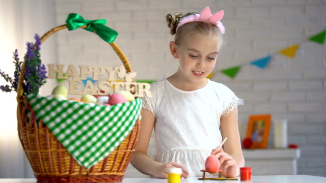 Creative-girl-in-white-dress-showing-painted-Easter-egg,-decorated-basket-table