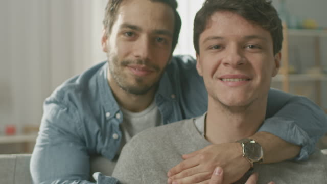 Portrait-of-a-Gentle-Male-Gay-Couple-at-Home.-Young-Man-Sits-on-a-Couch,-His-Partner-Embraces-him-from-Behind.-They-are-Happy-and-Smiling.-Room-Has-Modern-Interior.