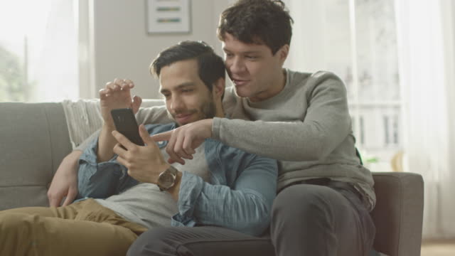 Cute-Male-Gay-Couple-Spend-Time-at-Home.-They-are-Lying-Down-on-a-Sofa-and-Use-a-Smartphone.-They-Browse-Online.-Partner's-Hand-is-Around-His-Lover.-They-Smile-and-Laugh.-Room-Has-Modern-Interior.
