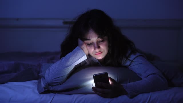 4K-breites-Video-von-einer-jungen-Frau,-die-im-Internet-plaudert-und-surft,-indem-sie-ihr-Smartphone-schläfrig,-gelangweilt-und-müde-bis-spät-in-die-Nacht-benutzt.-Dramatisches-dunkles-Licht.-Im-Internet,-Mobile-Sucht-und-Schlaflosigkeit-Konzept.