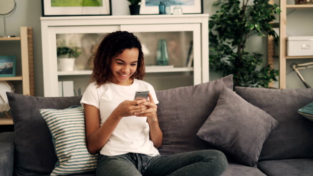 Schönes-afroamerikanisches-Mädchen-ist-mit-Smartphone-und-lachend-entspannt-auf-bequemen-Sofa-zu-Hause.-Moderne-Technik-und-Jugendleben-Konzept.